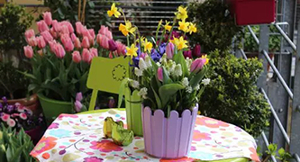 Blumen in Tontopf auf Tisch mit bemaltem Tuch, Gartenszene mit Tulpen, Narzissen und weiteren Pflanzen