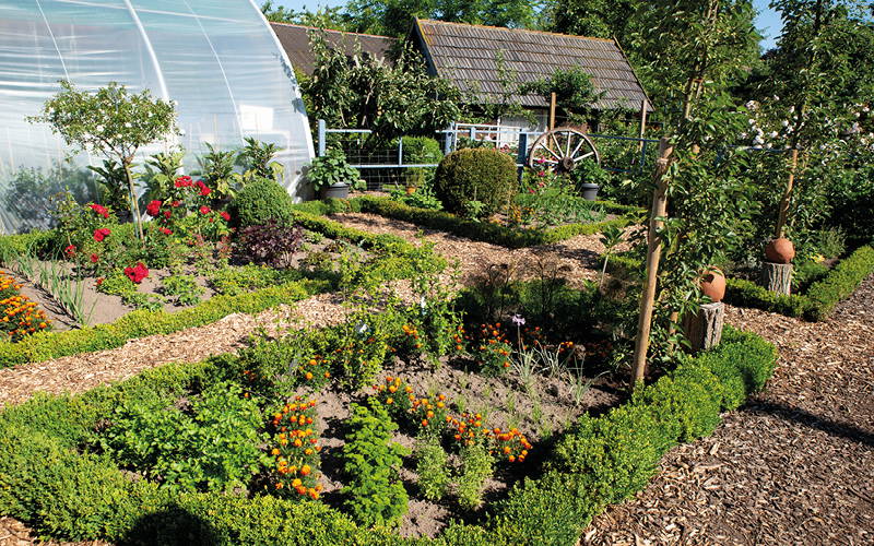 Garten mit bunten Blumen, Gemüsepflanzen, kleinen Bäumen und einer Scheune im Hintergrund.