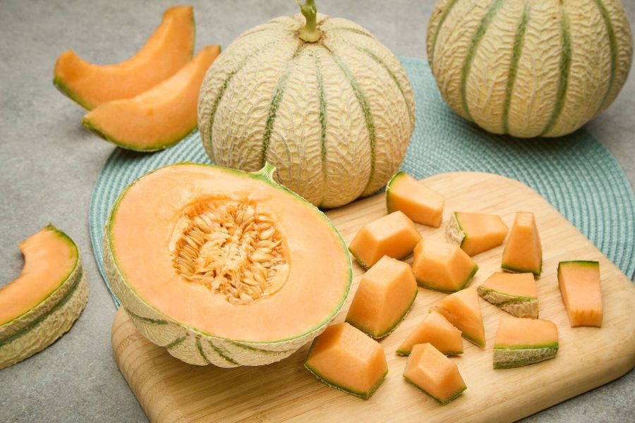Melone mit orangefarbenem Fruchtfleisch und mehreren Würfeln auf einem Holzbrett, daneben ganze und halbierte Melonen.
