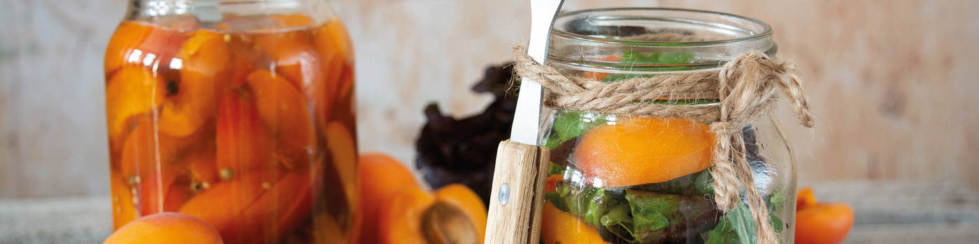 Gläser mit eingelegtem Obst und Gemüse, Orangen, und ein Messer auf Holzgriff.