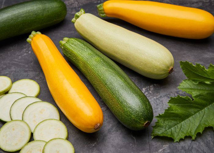 Verschiedene Zucchini in Grün, Gelb und Creme liegen auf einem dunklen Untergrund, einige Scheiben Zucchini und grüne Blätter daneben.