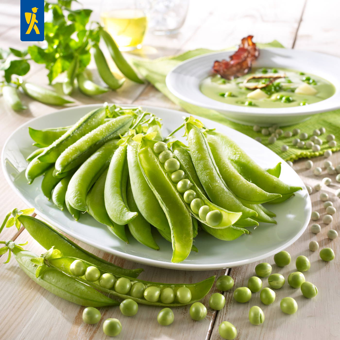 Grüne Erbsen in Hülsen, some Hülsen auf einem weißen Teller, im Hintergrund zwei Suppenschalen und ein Glas Wein.