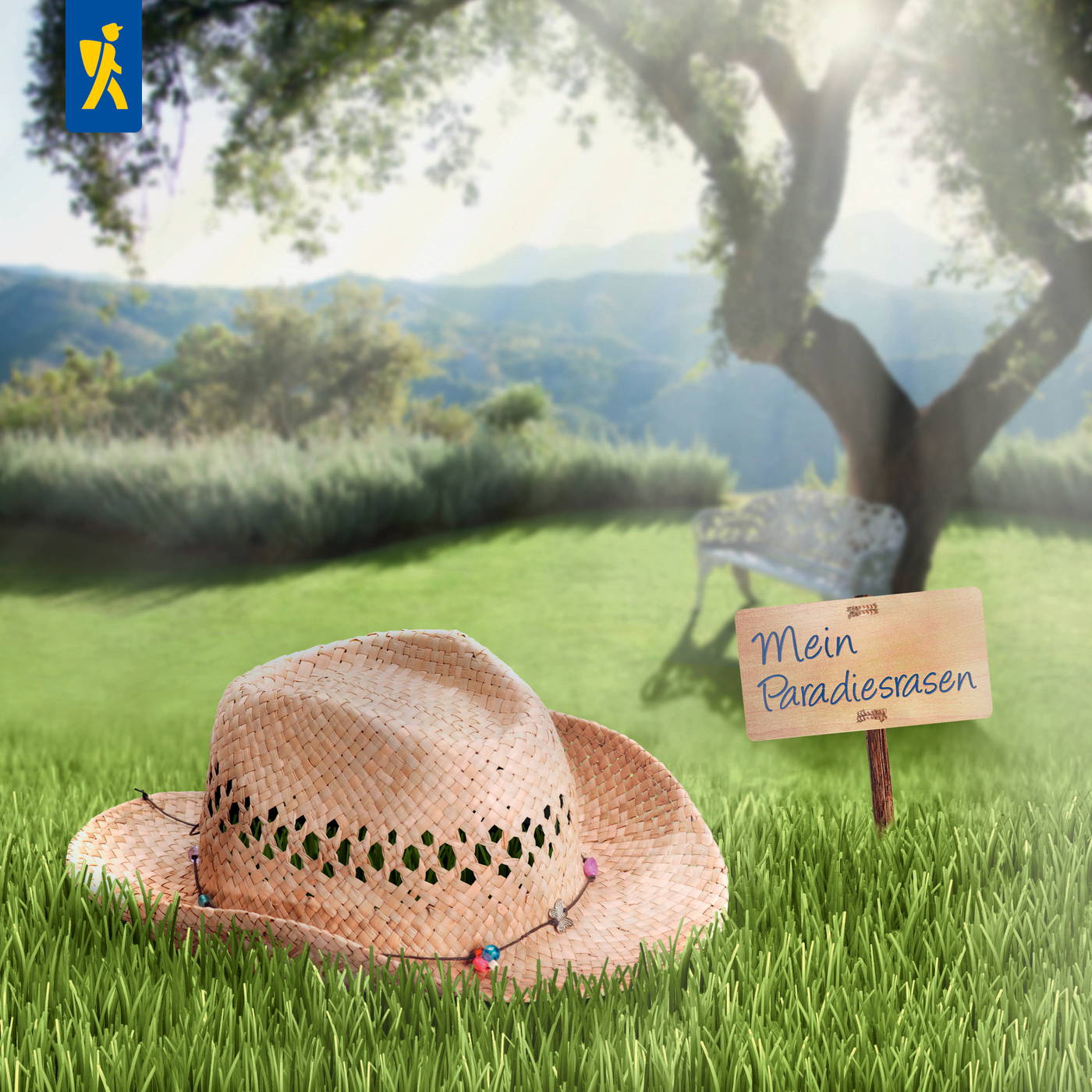 Hut auf grünem Rasen vor Baum mit Sitzbank im Hintergrund, kleines Schild mit "Mein Paradiesrasen".