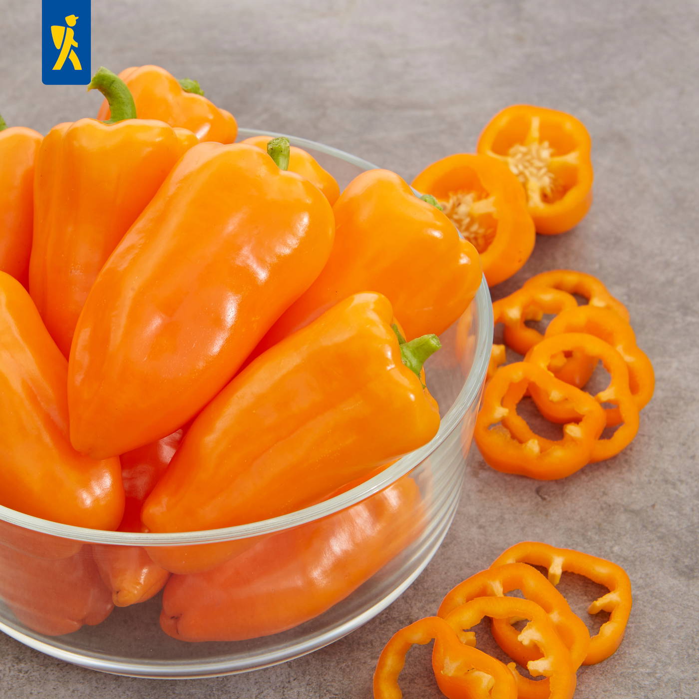 Orange Paprika in Schüssel, einige geschnittene Scheiben und Ringe daneben auf grauem Hintergrund.