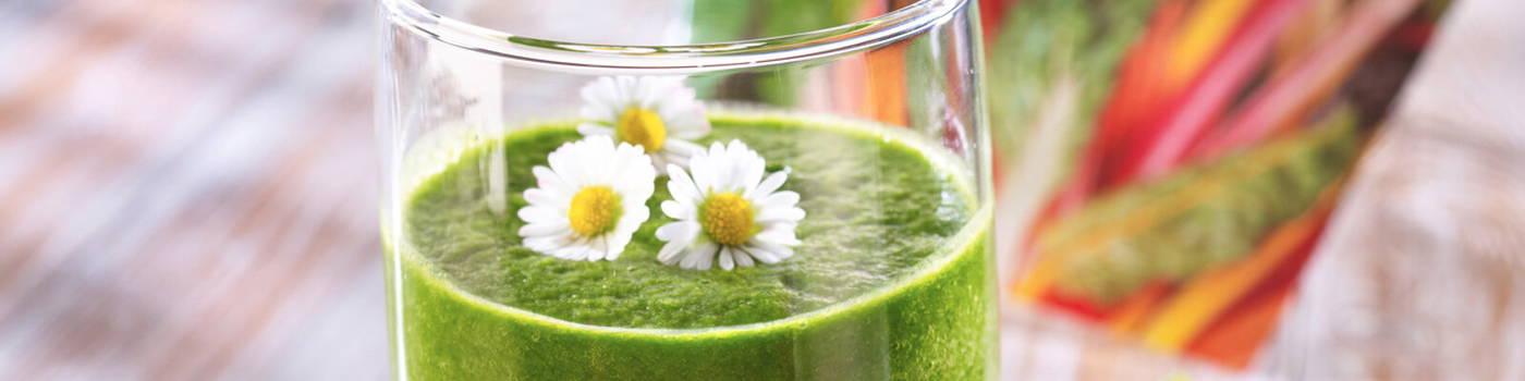 Grüner Smoothie im Glas mit Kamilleblumen und Strohhalm, im Hintergrund bunte Paprika.