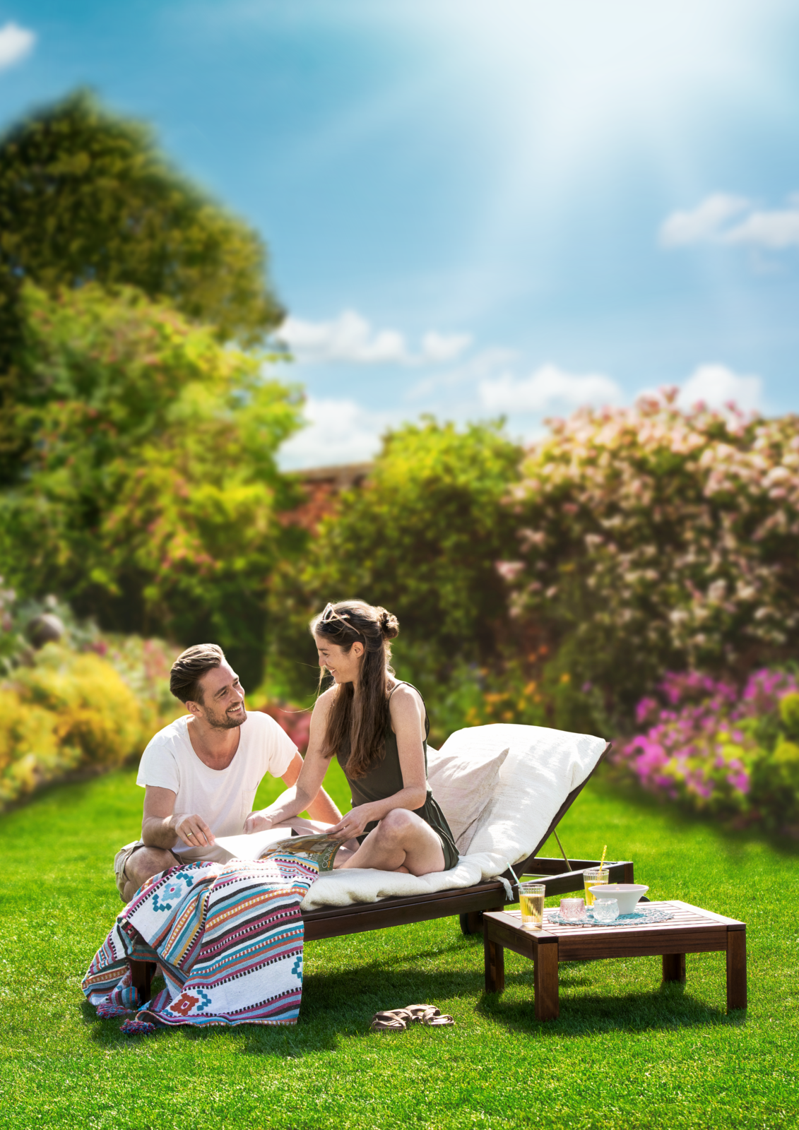 Zwei Menschen sitzen auf einer Liege im Garten auf Gras, lachen und schauen sich an, im Hintergrund blühende Bäume und Himmel.
