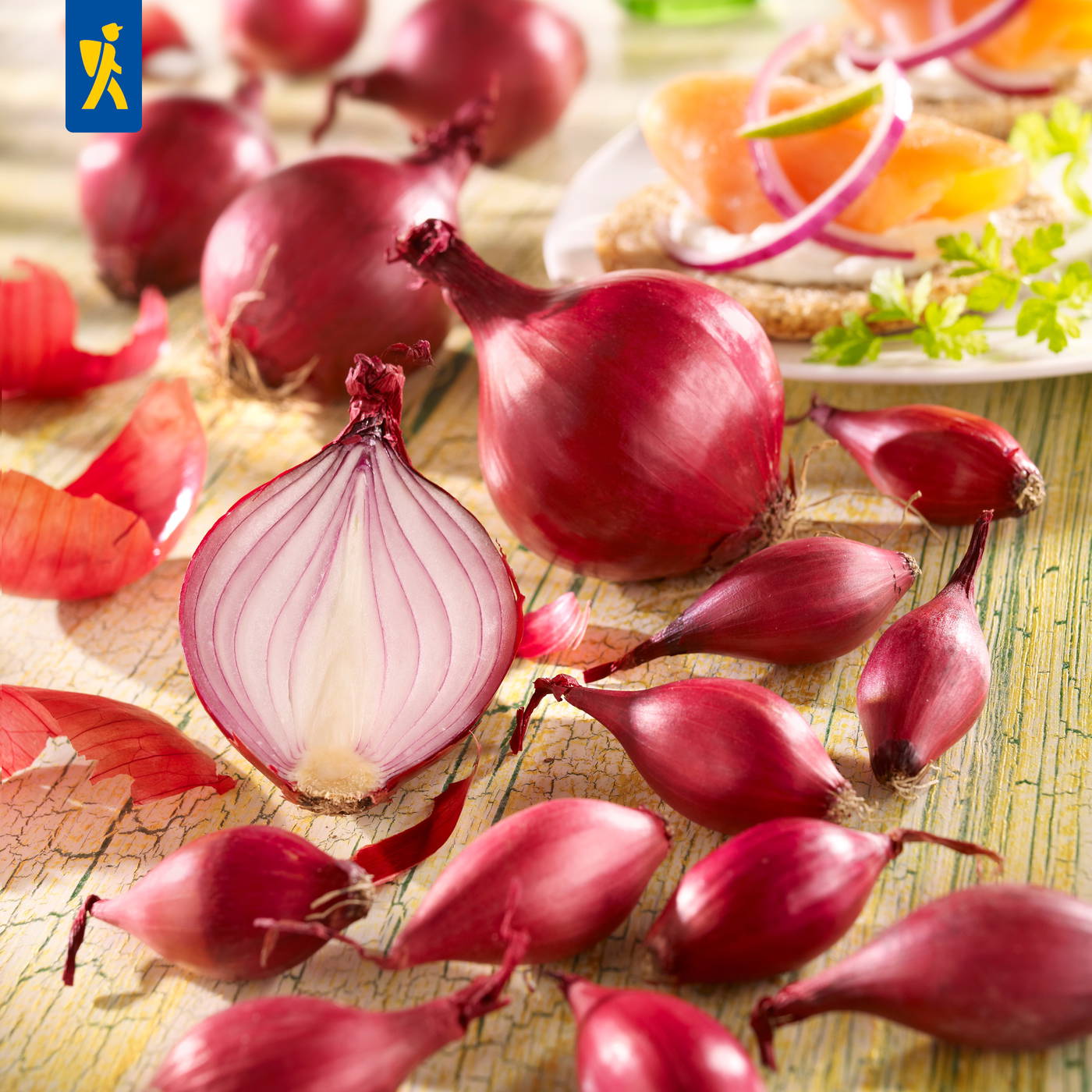 Rote Zwiebeln, halbiert und ganz, auf einem Holztablett mit einer belegten altrömischen Brotscheibe im Hintergrund.