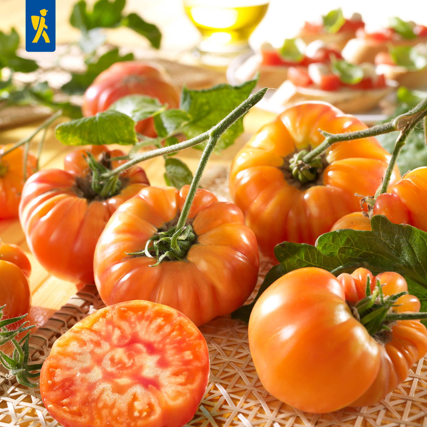 Rote Tomaten mit grünen Blättern auf einem Holztisch, im Hintergrund Tomatensalat und eine Flasche Öl.
