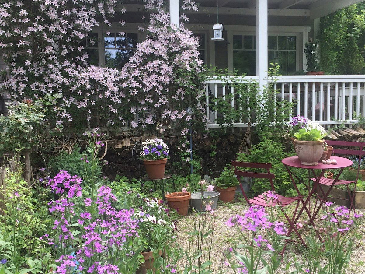 Garten mit pinken und lila Blumen, Gartenmöbeln und einem Haus im Hintergrund.