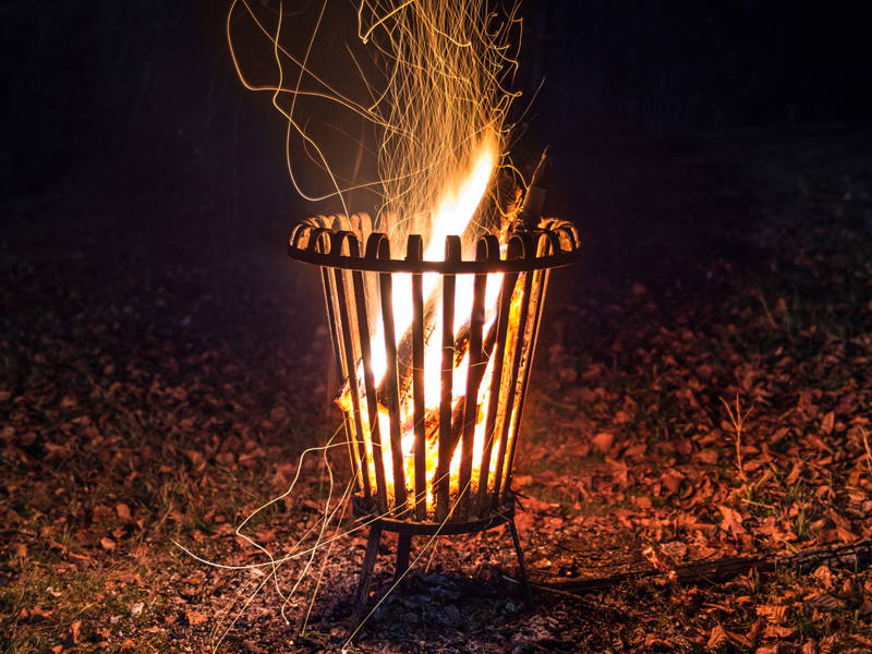 Feuer brennt in einem Metallkorb im Freien bei Nacht, Flammen und Funken steigen auf.