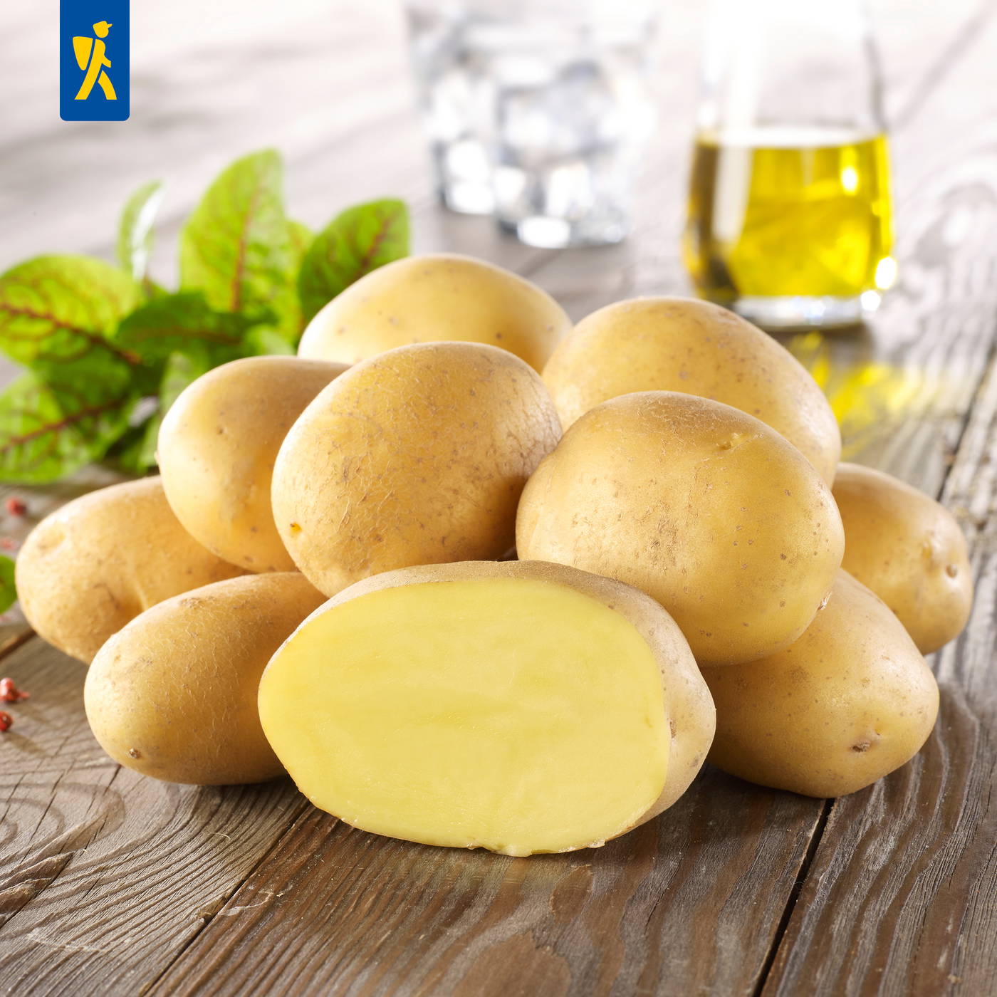 Mehrere rohe gelbe Kartoffeln auf einem Holztisch, eine Hälfte ist geschnitten, im Hintergrund ein Glas Öl und Blätter.