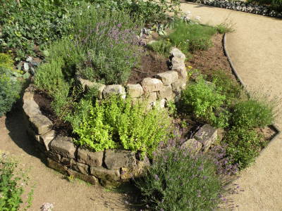 Kräuterspirale aus Natursteinen mit verschiedenen Kräutern in einem Garten neben einem Sandweg