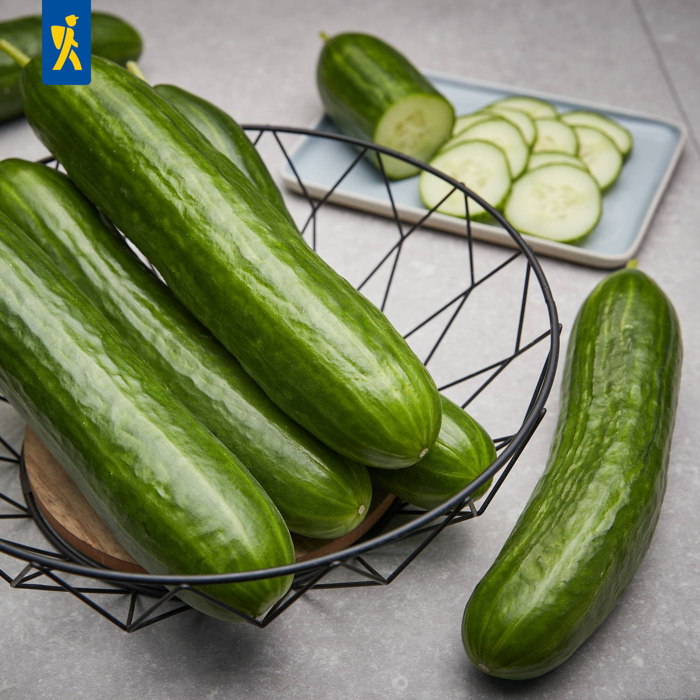Mehrere Salatgurken liegen im Drahtkorb auf grauer Oberfläche dabei liegen Gurkenscheiben auf einem Teller