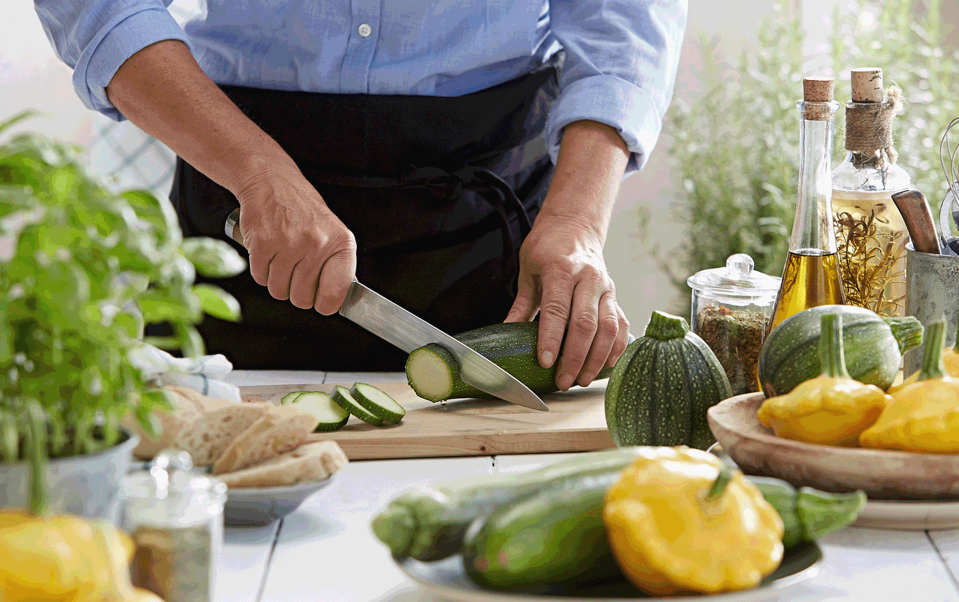Person schneidet Zucchini auf einem Küchenbrett, umgeben von Paprika, Zucchini, Kräutern und Ölflaschen.