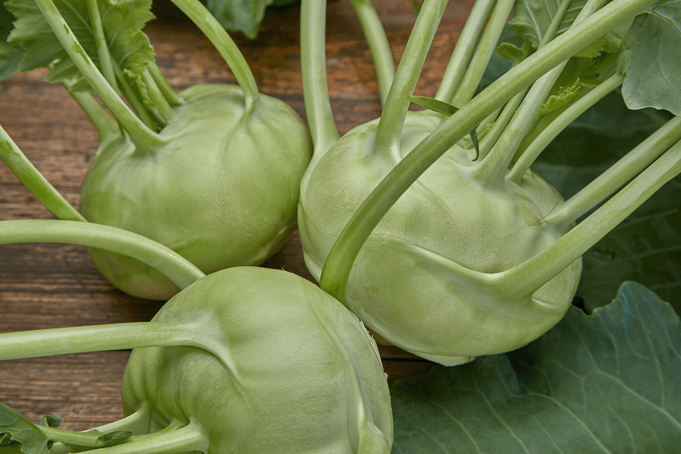 Drei grüne Kohlköpfe mit langen Stielen auf einer Holztischfläche.