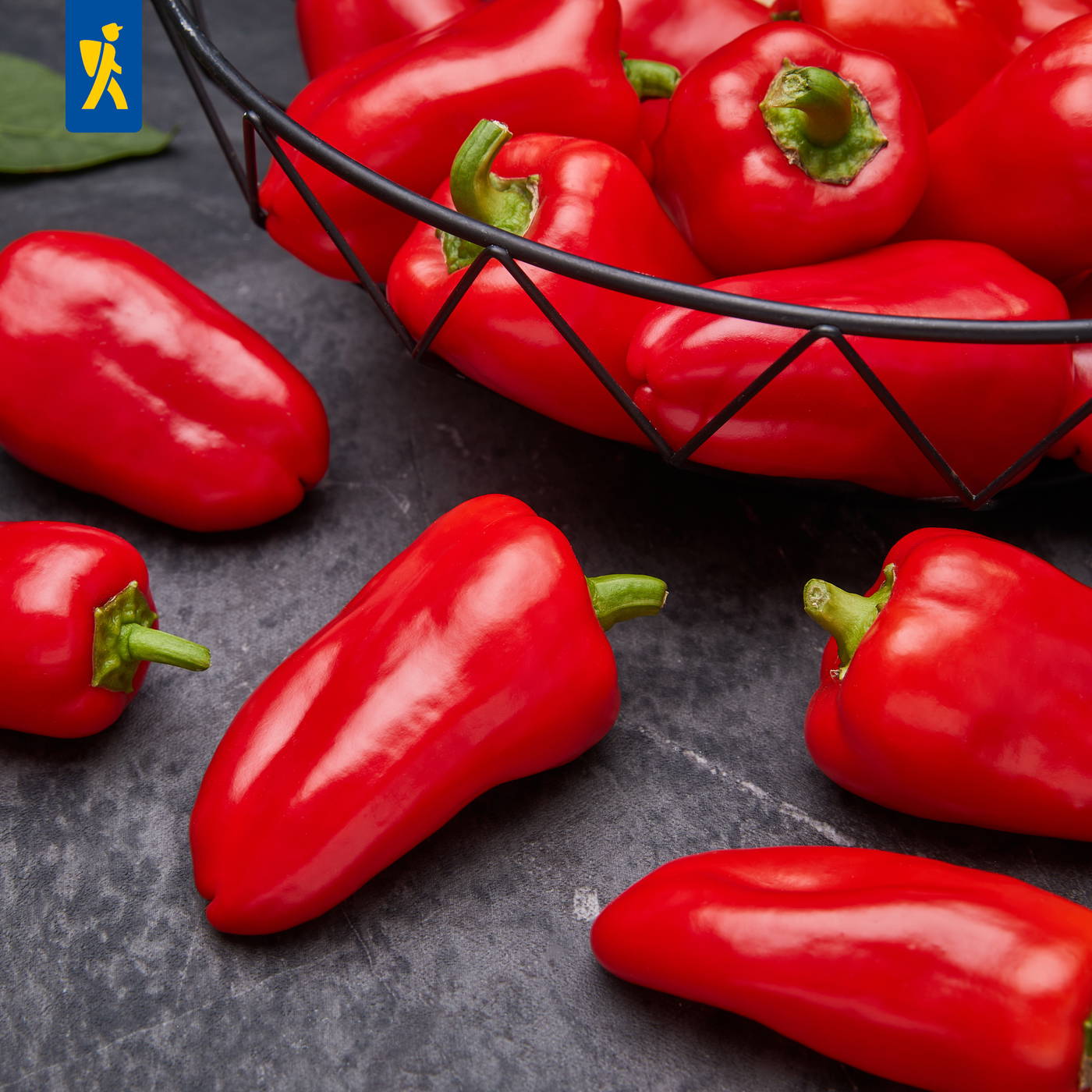 Rote Paprika liegen auf dunklem Steinboden, mehrere in einer schwarzen Gänseblümchenkorb, einige außerhalb.