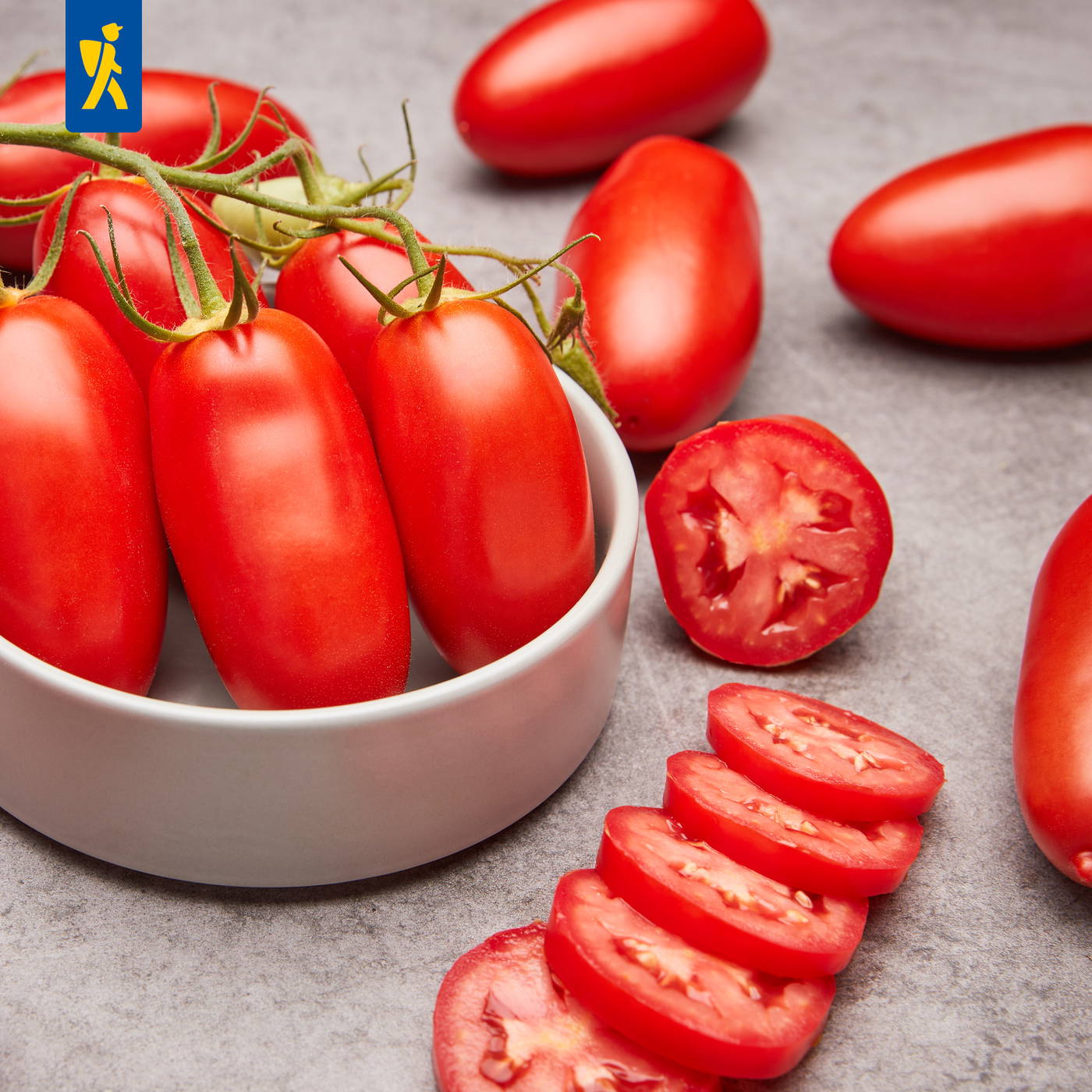 Rote Tomaten in einer weißen Schüssel, einige Tomaten halbiert, auf einem grauen Untergrund.