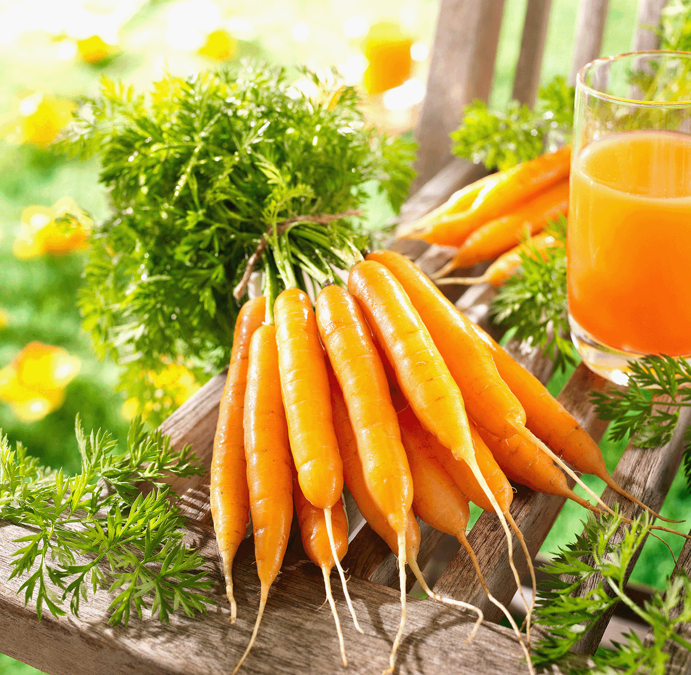 Bunte Karotten mit grünen Blättern auf Holzbrett, daneben Glas mit Orangensaft, im Hintergrund Blumen.
