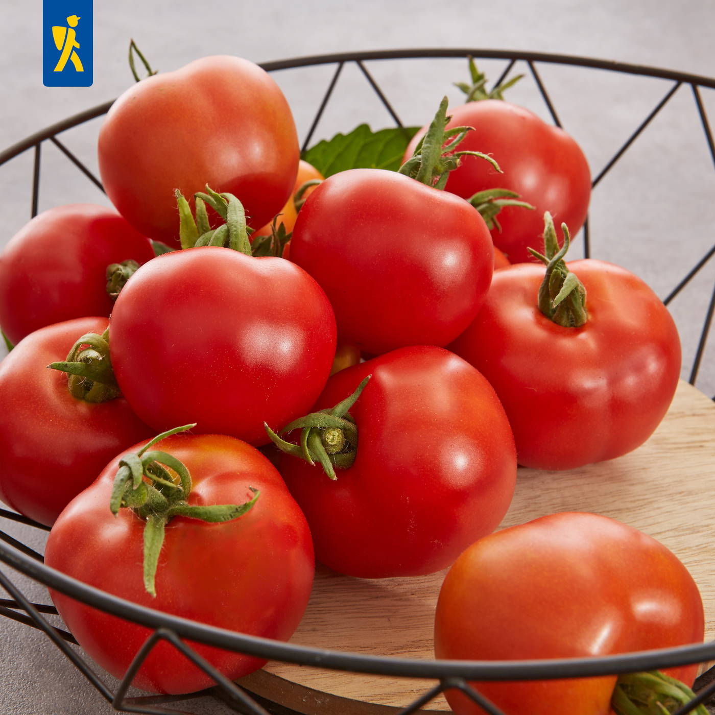 Rote Tomaten mit grünen Stielen in einer schwarzen Drahtkorb auf einem Holzbrett.