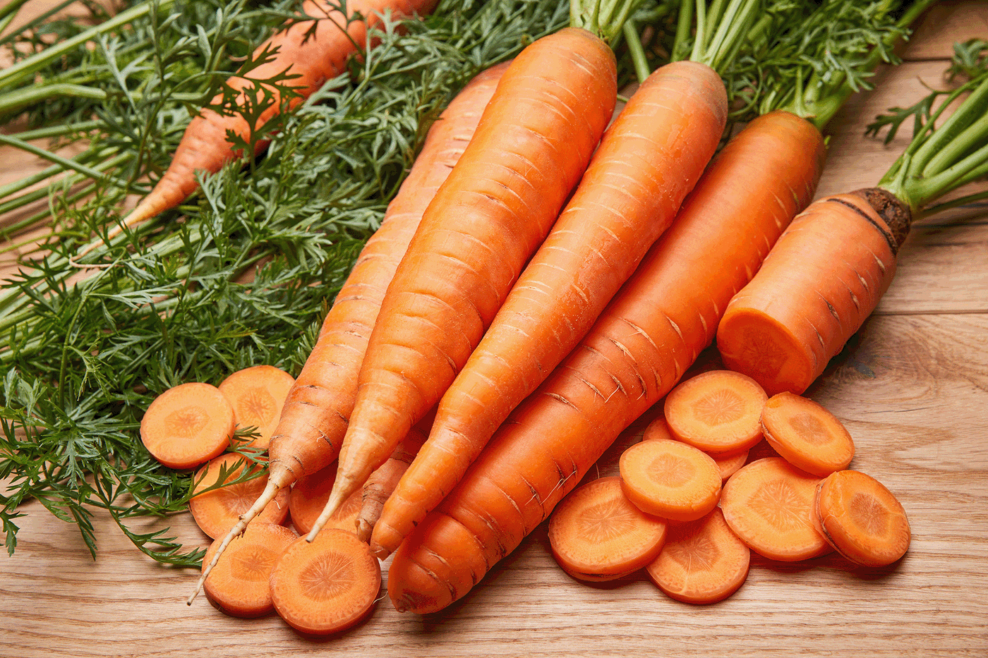 Frische Karotten mit grünen Blättern und geschnittenen Scheiben auf Holzuntergrund.