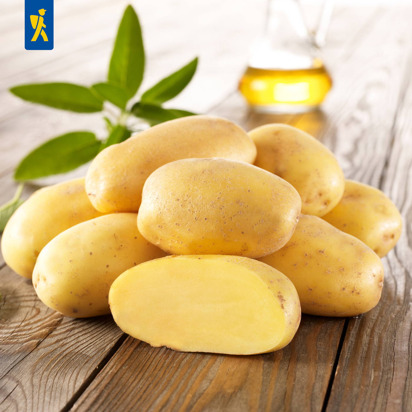 Mehrere gelbe Kartoffeln, eine davon halbiert, auf einem Holzbrett, mit grünen Blättern und einer Flasche Öl im Hintergrund.