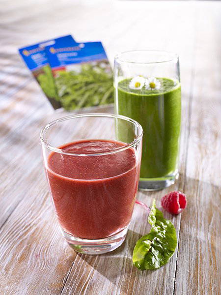 Zwei Gläser mit grünem und rotem Smoothie, Raspel und Verpackungen im Hintergrund auf Holzfläche.