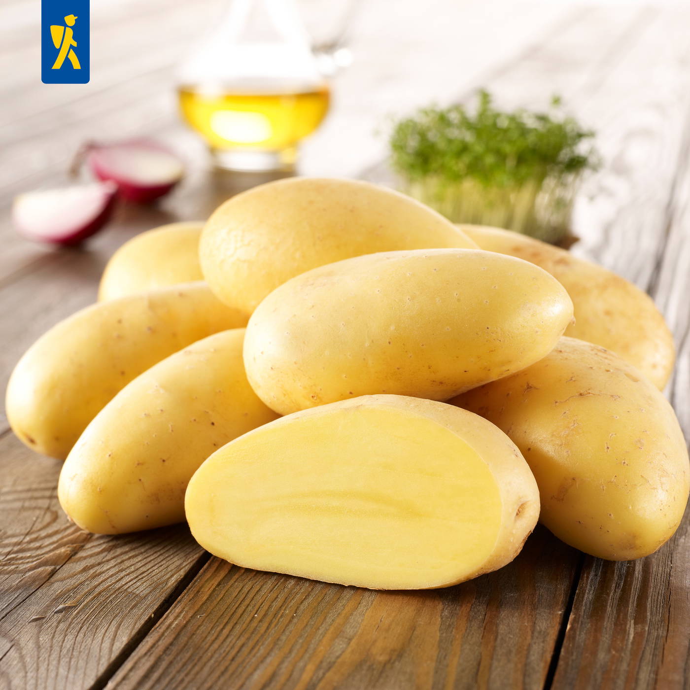 Stapel gelber Kartoffeln auf einem Holzbrett, im Hintergrund Radieschen, Öl und Kräuter.